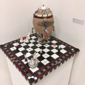 Image of a sculpture featuring a surreal head sitting on top of a chessboard surrounded by rabbits, toadstools and peg figures.